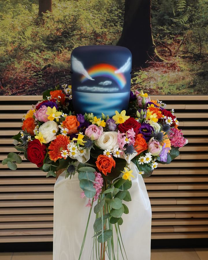 Urne mit Regenbogenmotiv auf buntem Blumenkranz