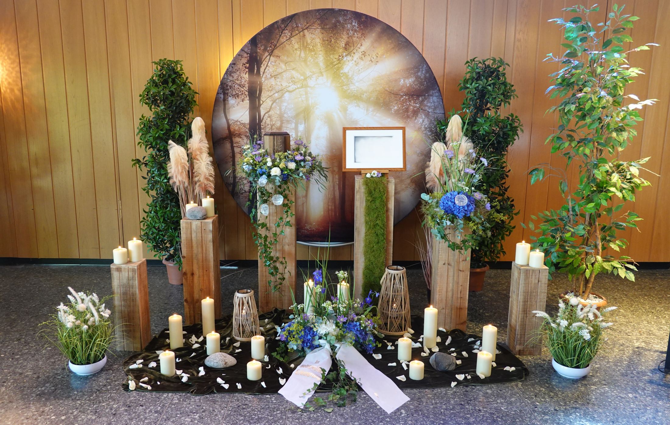 Trauerdekoration mit Kerzen, Blumen und Holzstelen vor einem Lichtmotiv
