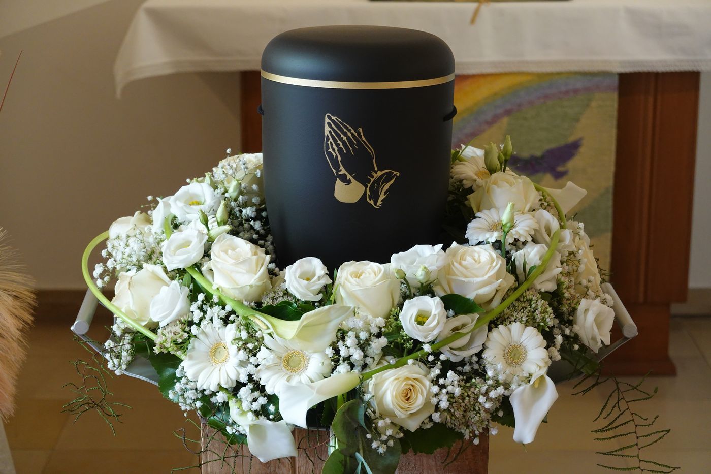 Schwarze Urne mit gefalteten Händen auf weißem Rosenkranz